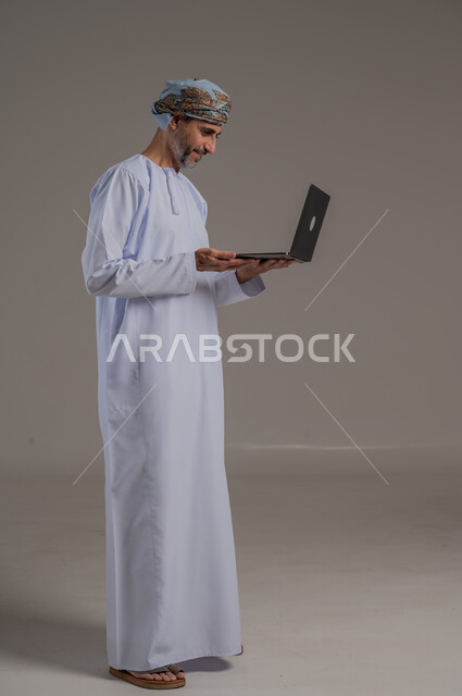 مفهوم تسهيل الاعمال والقدرة على إنجاز المهام عن بعد، استخدام التقنيات الحديثة والأجهزة المتطورة ، بورتريه لشاب عربي خليجي عماني يرتدي الدشداشة والعمامة يمسك بيده حاسوب محمول تظهر عليه علامات الفرح والسرور، الجسم بالطول الكامل، خلفية رمادية