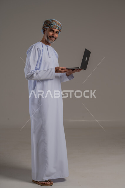 مفهوم تسهيل الاعمال والقدرة على إنجاز المهام عن بعد، استخدام التقنيات الحديثة والأجهزة المتطورة ، بورتريه لشاب عربي خليجي عماني يرتدي الدشداشة والعمامة يمسك بيده حاسوب محمول تظهر عليه علامات الفرح والسرور، الجسم بالطول الكامل، خلفية رمادية