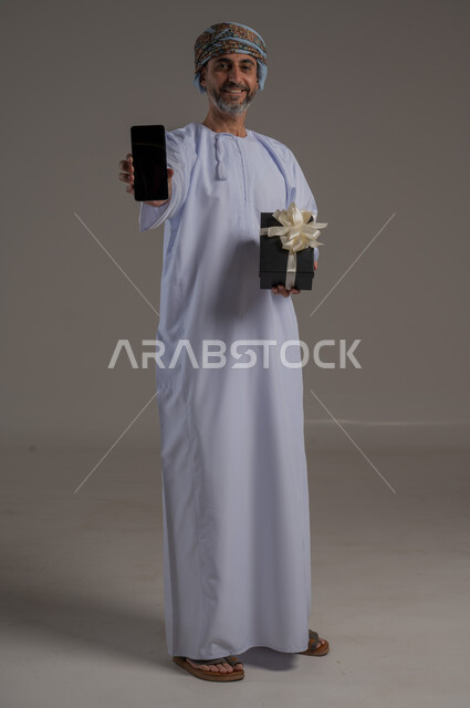 مفهوم تسهيل الاعمال والقدرة على إنجاز المهام عن بعد، استخدام التقنيات الحديثة والأجهزة المتطورة ، بورتريه لشاب عربي خليجي عماني يرتدي الدشداشة والعمامة يمسك بيده هاتف محمول تظهر عليه علامات الفرح والسرور، الجسم بالطول الكامل، خلفية رمادية