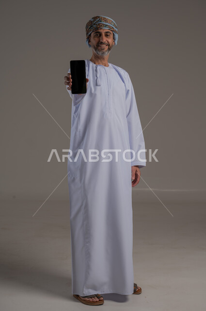 مفهوم تسهيل الاعمال والقدرة على إنجاز المهام عن بعد، استخدام التقنيات الحديثة والأجهزة المتطورة ، بورتريه لشاب عربي خليجي عماني يرتدي الدشداشة والعمامة يمسك بيده هاتف محمول تظهر عليه علامات الفرح والسرور، الجسم بالطول الكامل، خلفية رمادية