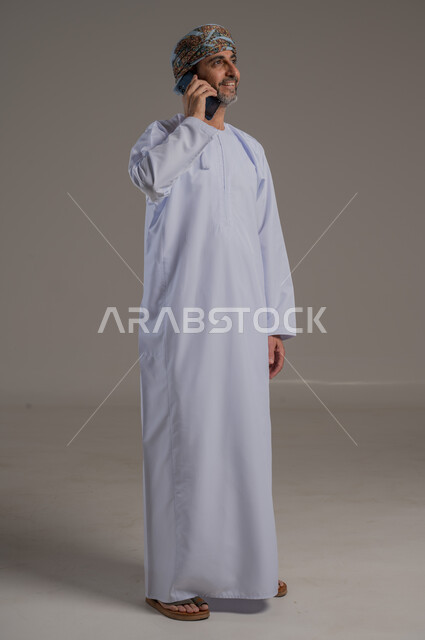 The concept of facilitating work and the ability to accomplish tasks remotely, using modern technologies and advanced devices, a portrait of a young Gulf Arab Omani man wearing a dishdasha and turban holding a mobile phone in his hand, showing signs of joy and happiness, full-length body, gray background