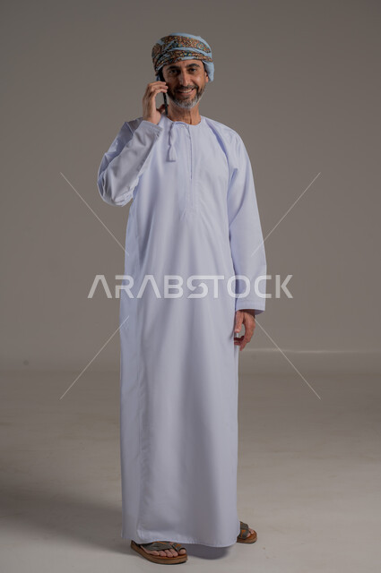 The concept of facilitating work and the ability to accomplish tasks remotely, using modern technologies and advanced devices, a portrait of a young Gulf Arab Omani man wearing a dishdasha and turban holding a mobile phone in his hand, showing signs of joy and happiness, full-length body, gray background