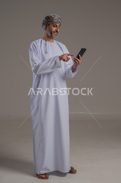 مفهوم تسهيل الاعمال والقدرة على إنجاز المهام عن بعد، استخدام التقنيات الحديثة والأجهزة المتطورة ، بورتريه لشاب عربي خليجي عماني يرتدي الدشداشة والعمامة يمسك بيده هاتف محمول تظهر عليه علامات الفرح والسرور، الجسم بالطول الكامل، خلفية رمادية