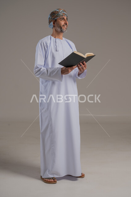الاجتهاد في العبادة، تلاوة وقراءة القرآن الكريم وتدبر معانيه، بورتريه لشاب عربي خليجي عماني يرتدي الدشداشة والعمامة يحمل بيده المصحف الشريف، الإكثار من ذكر الله تعالى، التقرب الى الله في شهر رمضان، الشعور بالخشوع والطمأنينة، الطول الكامل للجسم, خلفية رمادية