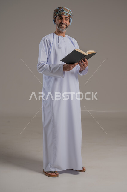 الاجتهاد في العبادة، تلاوة وقراءة القرآن الكريم وتدبر معانيه، بورتريه لشاب عربي خليجي عماني يرتدي الدشداشة والعمامة يحمل بيده المصحف الشريف، الإكثار من ذكر الله تعالى، التقرب الى الله في شهر رمضان، الشعور بالخشوع والطمأنينة، الطول الكامل للجسم, خلفية رمادية