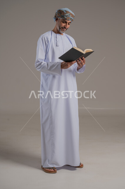 الاجتهاد في العبادة، تلاوة وقراءة القرآن الكريم وتدبر معانيه، بورتريه لشاب عربي خليجي عماني يرتدي الدشداشة والعمامة يحمل بيده المصحف الشريف، الإكثار من ذكر الله تعالى، التقرب الى الله في شهر رمضان، الشعور بالخشوع والطمأنينة، الطول الكامل للجسم, خلفية رمادية