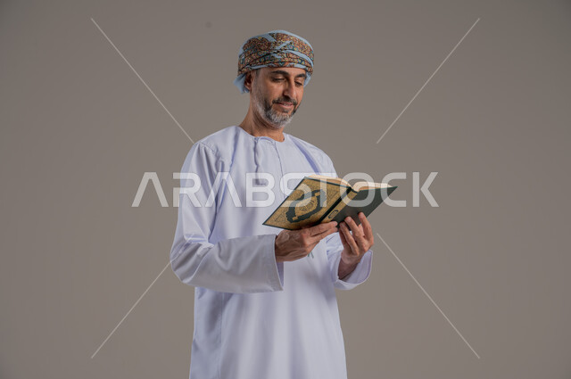 الاجتهاد في العبادة، تلاوة وقراءة القرآن الكريم وتدبر معانيه، بورتريه لشاب عربي خليجي عماني يرتدي الدشداشة والعمامة يحمل بيده المصحف الشريف، الإكثار من ذكر الله تعالى، التقرب الى الله في شهر رمضان، الشعور بالخشوع والطمأنينة، الطول الكامل للجسم, خلفية رمادية