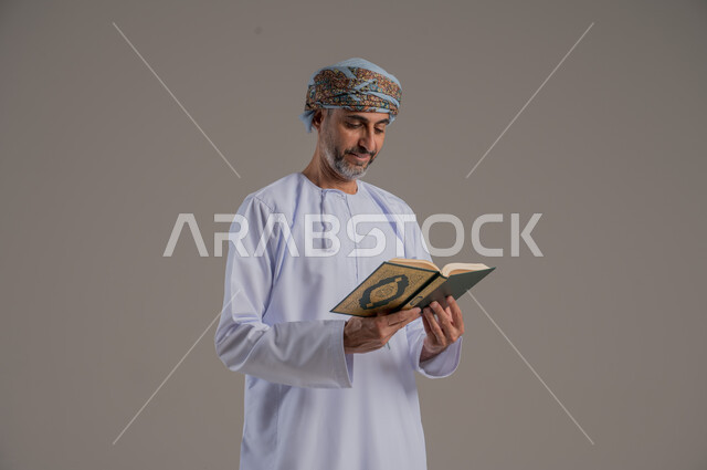 الاجتهاد في العبادة، تلاوة وقراءة القرآن الكريم وتدبر معانيه، بورتريه لشاب عربي خليجي عماني يرتدي الدشداشة والعمامة يحمل بيده المصحف الشريف، الإكثار من ذكر الله تعالى، التقرب الى الله في شهر رمضان، الشعور بالخشوع والطمأنينة، الطول الكامل للجسم, خلفية رمادية