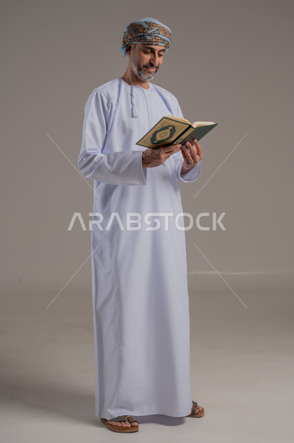 الاجتهاد في العبادة، تلاوة وقراءة القرآن الكريم وتدبر معانيه، بورتريه لشاب عربي خليجي عماني يرتدي الدشداشة والعمامة يحمل بيده المصحف الشريف، الإكثار من ذكر الله تعالى، التقرب الى الله في شهر رمضان، الشعور بالخشوع والطمأنينة، الطول الكامل للجسم, خلفية رمادية