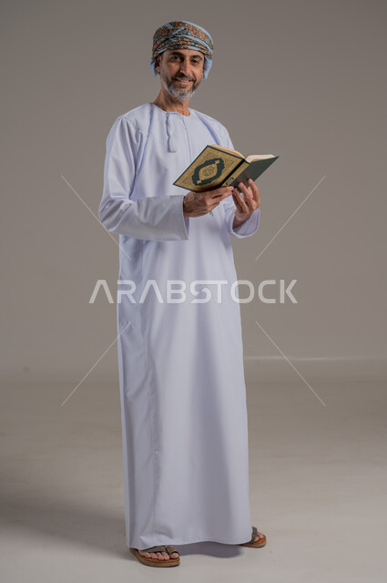 الاجتهاد في العبادة، تلاوة وقراءة القرآن الكريم وتدبر معانيه، بورتريه لشاب عربي خليجي عماني يرتدي الدشداشة والعمامة يحمل بيده المصحف الشريف، الإكثار من ذكر الله تعالى، التقرب الى الله في شهر رمضان، الشعور بالخشوع والطمأنينة، الطول الكامل للجسم, خلفية رمادية