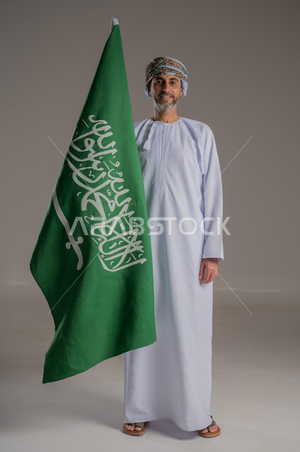Pride and honor in the nation's achievements, the slogan of national identity and the banner of Islam and monotheism, a portrait of a smiling Gulf Arab Omani man wearing a dishdasha and turban holding the founding flag in his hand, the anniversary of the founding of the first Saudi state, February 22, gray background