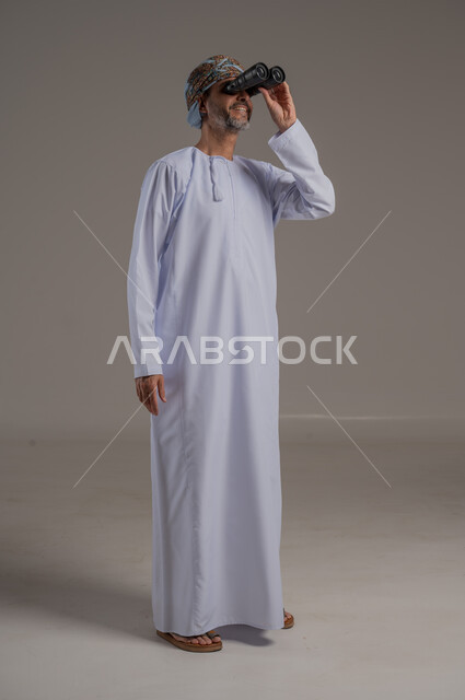 Seeing distant objects through binoculars, love of adventure and exploration, portrait of an Arab Gulf student wearing a dishdasha and turban looking through the perspective with different facial gestures, future planning for goals and ambitions, full-length body image, gray background