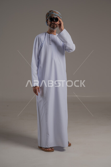 رؤية الاشياء البعيدة عبر المنظار، حب المغامرة والاستكشاف، بورتريه لطالب عربي خليجي يرتدي الدشداشة والعمامة  ينظر من خلال المنظور بإيماءات وجه مختلفة, التخطيط المستقبلي للأهداف والطموحات، صورة الجسم بالطول الكامل، خلفية رمادية