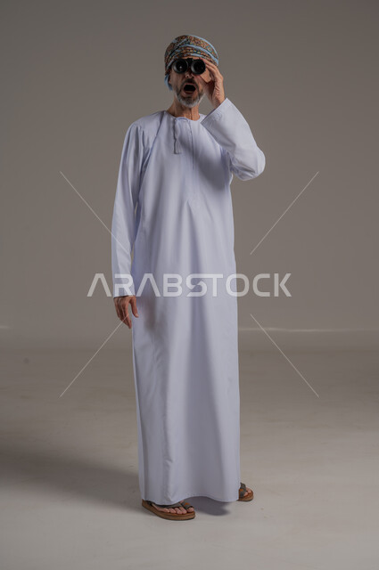 رؤية الاشياء البعيدة عبر المنظار، حب المغامرة والاستكشاف، بورتريه لطالب عربي خليجي يرتدي الدشداشة والعمامة  ينظر من خلال المنظور بإيماءات وجه مختلفة, التخطيط المستقبلي للأهداف والطموحات، صورة الجسم بالطول الكامل، خلفية رمادية