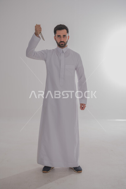 تعابير العبوس والانزعاج، نظرات التحدي والغضب، بورتريه لرجل عربي خليجي سعودي يرتدي الثوب التقليدي يحمل بيده سكينه يوجهها نحو رأسه ينظر الى الكاميرا بثقة، مفهوم عدم القبول والرضا، صورة الجسم بالطول الكامل، خلفية بيضاء 