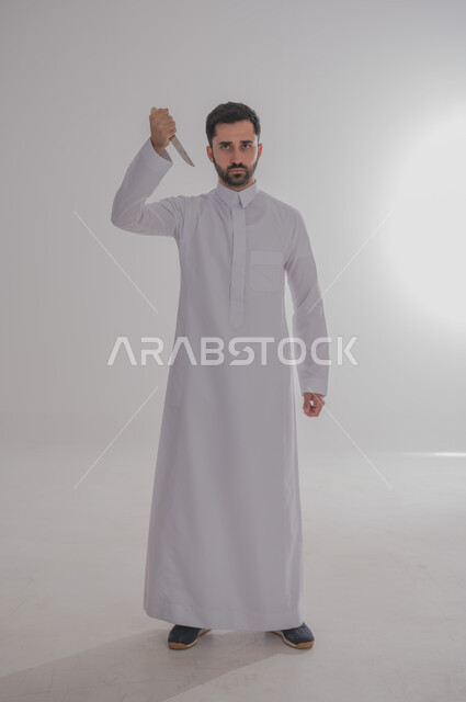 تعابير العبوس والانزعاج، نظرات التحدي والغضب، بورتريه لرجل عربي خليجي سعودي يرتدي الثوب التقليدي يحمل بيده سكينه يوجهها نحو رأسه ينظر الى الكاميرا بثقة، مفهوم عدم القبول والرضا، صورة الجسم بالطول الكامل، خلفية بيضاء 