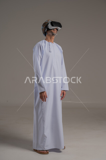دمج التقنيات والتكنولوجيا الحديثة مع المتعة والتسلية، محاكاة العالم الخيالي، بورتريه لرجل عربي خليجي عماني يرتدي دشداشة وعمامة ونظارات الواقع الافتراضي ثلاثية الأبعاد، ايماءات الاندماج والتركيز، صورة الجسم بالطول الكامل، خلفية رمادية