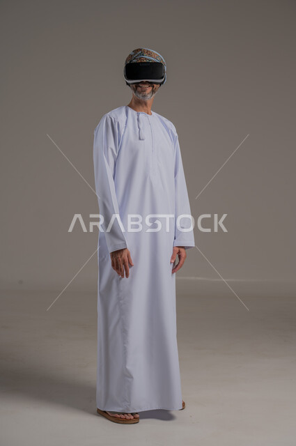 دمج التقنيات والتكنولوجيا الحديثة مع المتعة والتسلية، محاكاة العالم الخيالي، بورتريه لرجل عربي خليجي عماني يرتدي دشداشة وعمامة ونظارات الواقع الافتراضي ثلاثية الأبعاد، ايماءات الاندماج والتركيز، صورة الجسم بالطول الكامل، خلفية رمادية