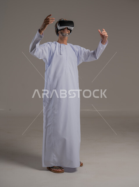 Merging modern technology and techniques with fun and entertainment, simulating the imaginary world, portrait of an Arab Gulf Omani man wearing a dishdasha, turban and 3D virtual reality glasses, gestures of integration and focus, full-length body image, gray background