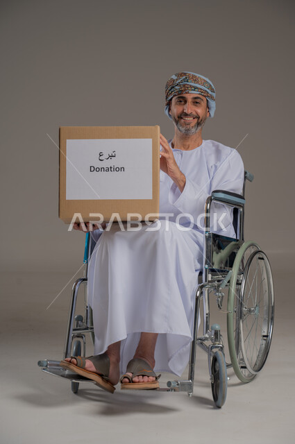 مفهوم التبرع والأعمال الخيرية الإسلاميه في سلطنه عمان، بورتريه لرجل عربي عماني خليجي من ذوي الاحتياجات الخاصة يرتدي الدشداشة والعمامة يجلس على كرسي يمسك بيده صندوق كرتوني، النظر إلى الكاميرا بإيماءات السرور، خلفية رمادية
