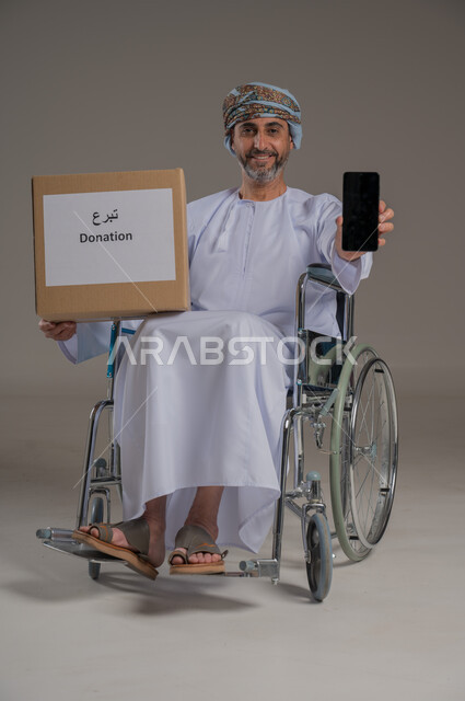 مفهوم التبرع والأعمال الخيرية الإسلاميه في سلطنه عمان، بورتريه لرجل عربي عماني خليجي من ذوي الاحتياجات الخاصة يرتدي الدشداشة والعمامة يجلس على كرسي يمسك بيده صندوق كرتوني، النظر إلى الكاميرا بإيماءات السرور، خلفية رمادية