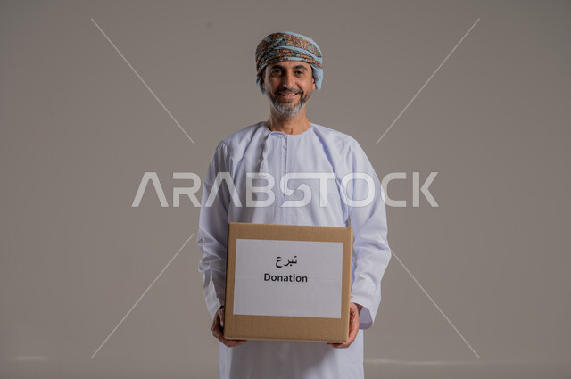 مفهوم التبرع والأعمال الخيرية الإسلاميه في سلطنه عمان، بورتريه لرجل عربي عماني خليجي يرتدي الدشداشة والعمامة يمسك بيده صندوق كرتوني، النظر إلى الكاميرا بإيماءات السرور، خلفية رمادية