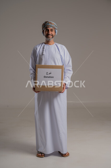 The concept of donation and Islamic charity in the Sultanate of Oman, portrait of an Arab Omani Gulf man wearing a dishdasha and turban holding a cardboard box in his hand, looking at the camera with gestures of pleasure, gray background