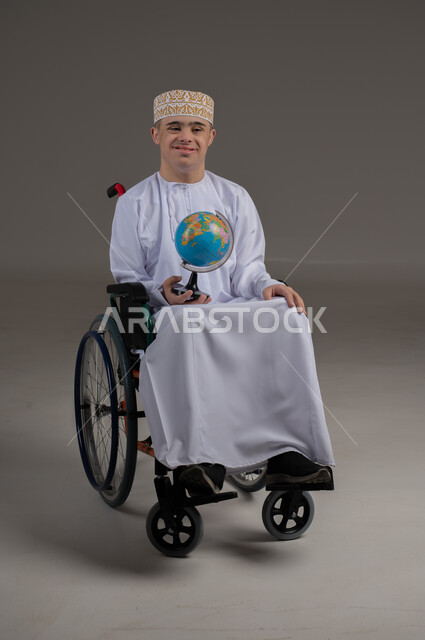 People with special needs and determination, looking at the camera with expressions of happiness and pleasure, portrait of a young Arab Gulf Omani man wearing a kummah with a dishdasha and sitting in a wheelchair holding a globe in his hand, gray background
