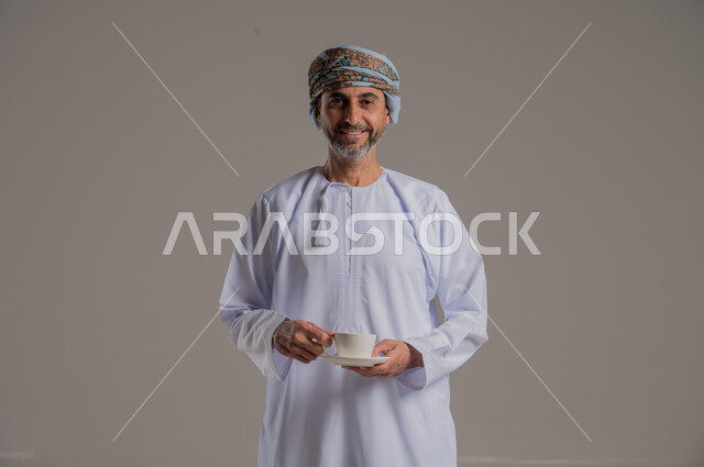 التمسك بالقيم الكريمة المتوارثة من الأجداد، بورتريه لرجل عربي خليجي عماني يرتدي العمامة والدشداشة يحمل بيده فنجان من القهوة، الاستمتاع بالمشروبات الساخنة، خلفية رمادية
