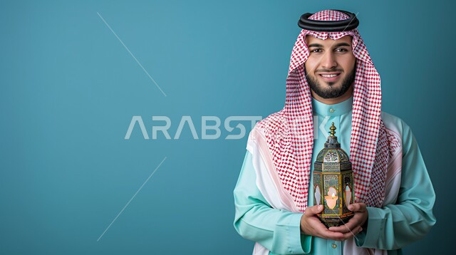 The concept of masculinity, elegance and elegance, attention to external appearance, a Saudi Gulf Arab man wearing a thobe with a shemagh, holding a Ramadan lantern in his hands and looking at the camera with gestures of self-confidence and pleasure, elegant Saudi fashion, blue background