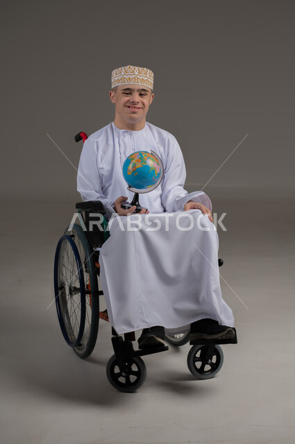 القيام برحلة سياحية, مفهوم البحث والاستكشاف حول العالم, بورتريه لشاب عربي خليجي عماني مقعد مصاب بمتلازمة داون يرتدي الدشداشة يجلس على كرسي متحرك يحمل بيده مجسم الكرة الارضية, صورة الجسم بالطول الكامل, خلفية رمادية