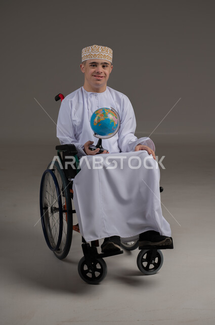 القيام برحلة سياحية, مفهوم البحث والاستكشاف حول العالم, بورتريه لشاب عربي خليجي عماني مقعد مصاب بمتلازمة داون يرتدي الدشداشة يجلس على كرسي متحرك يحمل بيده مجسم الكرة الارضية, صورة الجسم بالطول الكامل, خلفية رمادية