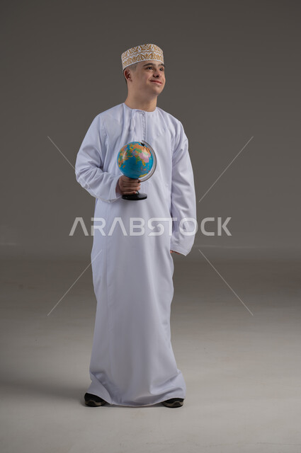 القيام برحلة سياحية, مفهوم البحث والاستكشاف حول العالم, بورتريه لشاب عربي خليجي عماني من ذوي الاحتياجات الخاصة يرتدي الدشداشة والكمة يحمل بيده مجسم الكرة الارضية, صورة الجسم بالطول الكامل, خلفية رمادية