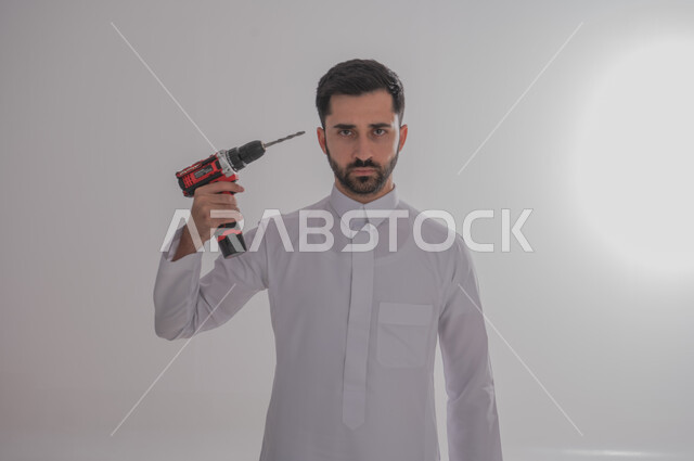 فني بناء وكسوة، لقطة مقربة لرجل خليجي عربي يرتدي ثوبًا تقليديًا ويحمل مطرقة ويعمل في قطاع الهندسة، وينظر إلى الكاميرا بإيماءات غاضبة، مهن ووظائف الشباب السعودي، خدمات الأعمال والصيانة، خلفية رمادية