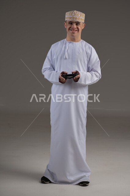 قضاء أوقات الفراغ باللعب بألعاب البلايستيشن الترفيهية ، إيماءات السعادة والسرور, تقنيات حديثة مسلية, بورتريه لطفل عماني من ذوي الاحتياجات الخاصة يرتدي الدشاشة والكمة يحمل بيده عصا التحكم، صورة الجسم بالطول الكامل، خلفية رمادية