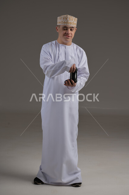 قضاء أوقات الفراغ باللعب بألعاب البلايستيشن الترفيهية ، إيماءات السعادة والسرور, تقنيات حديثة مسلية, بورتريه لطفل عماني من ذوي الاحتياجات الخاصة يرتدي الدشاشة والكمة يحمل بيده عصا التحكم، صورة الجسم بالطول الكامل، خلفية رمادية