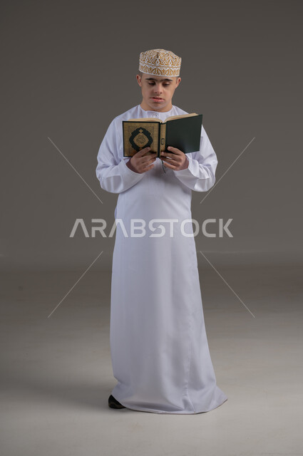 حفظ وتلاوة القرآن الكريم، ايماءات السعادة والسرور، تعلم الطقوس الدينية، بورتريه لطفل عماني من ذوي الاحتياجات الخاصة يرتدي الدشاشة والكمة يحمل بيده المصحف الشريف، العبادة والتقرب إلى الله، صورة الجسم بالطول الكامل، خلفية رمادية