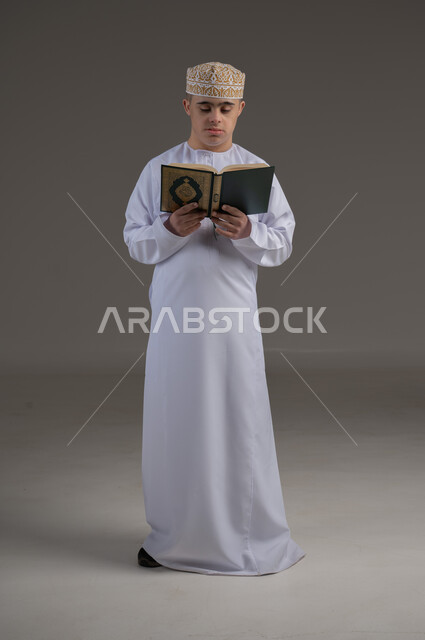 Memorizing and reciting the Holy Quran, gestures of happiness and pleasure, learning religious rituals, portrait of a special needs Omani child wearing a dishdasha and a kummah holding the Holy Quran in his hand, worship and getting closer to God, full body image, gray background