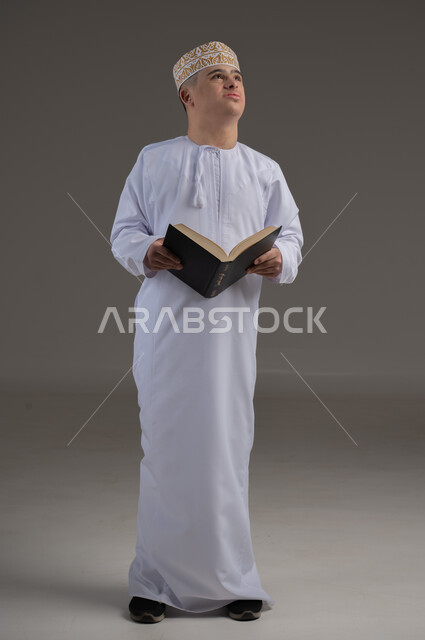 حفظ وتلاوة القرآن الكريم، ايماءات السعادة والسرور، تعلم الطقوس الدينية، بورتريه لطفل عماني من ذوي الاحتياجات الخاصة يرتدي الدشاشة والكمة يحمل بيده المصحف الشريف، العبادة والتقرب إلى الله، صورة الجسم بالطول الكامل، خلفية رمادية