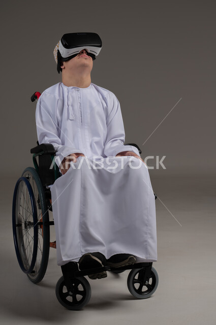 Simulating the imaginary world and interacting with the movie, improving the quality of life for people with motor disabilities, using wheelchairs to help with movement and mobility, portrait of an Arab Gulf Omani man wearing a kimmah and a dishdasha watching movies through 3D virtual reality glasses, gray background