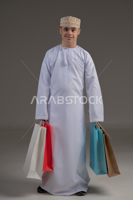 ذوي الإحتياجات الخاصة والهمم, بورتريه لشاب عربي خليجي عماني يرتدي الكمه والدشداشة يحمل بيده مجموعة من الحقائب الملونة، الاعاقة الحركية وحمل اكياس التسوق، مفهوم الترفيه والتبضع, صورة الجسم بالطول الكامل، خلفية رمادية