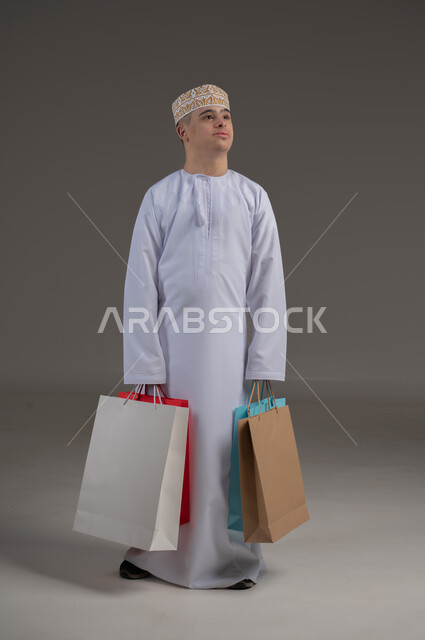 ذوي الإحتياجات الخاصة والهمم, بورتريه لشاب عربي خليجي عماني يرتدي الكمه والدشداشة يحمل بيده مجموعة من الحقائب الملونة، الاعاقة الحركية وحمل اكياس التسوق، مفهوم الترفيه والتبضع, صورة الجسم بالطول الكامل، خلفية رمادية