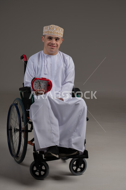 التذكيرات والتنبيهات الثابته، الدقة في المواعيد، بورتريه لرجل عربي خليجي عماني من ذوي الاحتياجات الخاصة يرتدي الدشاشة والكمة يجلس على كرسي متكرك يحمل بيده ساعة رقمية صغيرة, إنجاز المهامات في الوقت، ايماءات السعادة والسرور، خلفية رمادية