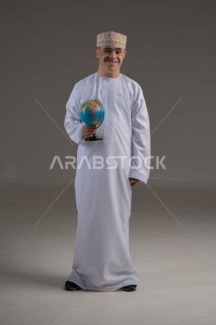 القيام برحلة سياحية, مفهوم البحث والاستكشاف حول العالم, بورتريه لشاب عربي خليجي عماني من ذوي الاحتياجات الخاصة يرتدي الدشداشة والكمة يحمل بيده مجسم الكرة الارضية, صورة الجسم بالطول الكامل, خلفية رمادية