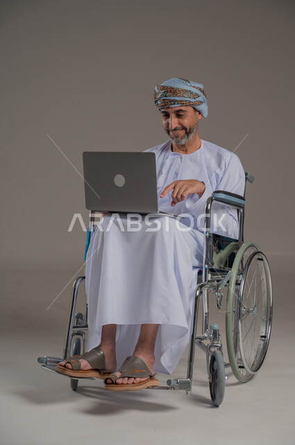 ذوي الإحتياجات الخاصة والهمم, مفهوم اللعب والتسلية, بورتريه لشاب عربي خليجي عماني يرتدي العمامة والدشداشة يجلس على كرسي متحرك ويحمل بيديه جهاز اللابتوب, صورة الجسم بالطول الكامل، خلفية رمادية