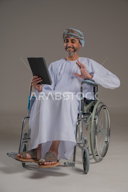 People with special needs and determination, the concept of play and entertainment, a portrait of a young Arab Gulf Omani man wearing a turban and a dishdasha sitting in a wheelchair and holding a tablet in his hands, a full-length body image, gray background