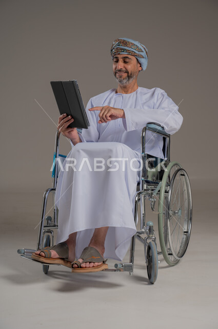ذوي الإحتياجات الخاصة والهمم, مفهوم اللعب والتسلية, بورتريه لشاب عربي خليجي عماني يرتدي العمامة والدشداشة يجلس على كرسي متحرك ويحمل بيديه جهاز تابليت, صورة الجسم بالطول الكامل، خلفية رمادية