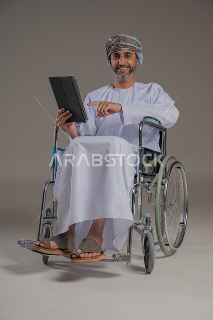 ذوي الإحتياجات الخاصة والهمم, مفهوم اللعب والتسلية, بورتريه لشاب عربي خليجي عماني يرتدي العمامة والدشداشة يجلس على كرسي متحرك ويحمل بيديه جهاز تابليت, صورة الجسم بالطول الكامل، خلفية رمادية