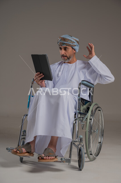 ذوي الإحتياجات الخاصة والهمم, مفهوم اللعب والتسلية, بورتريه لشاب عربي خليجي عماني يرتدي العمامة والدشداشة يجلس على كرسي متحرك ويحمل بيديه جهاز تابليت, صورة الجسم بالطول الكامل، خلفية رمادية