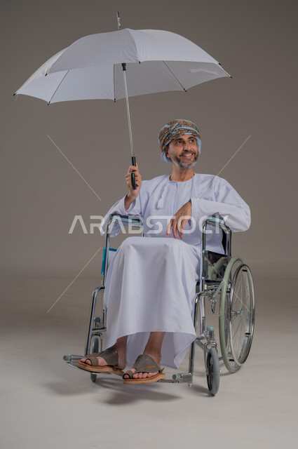 الحماية من الأمطار في الشتاء، دمج ذوي الهمم بالمجتمع العماني، التظلل من أشعة الشمس، بورتريه لرجل عربي خليجي عماني مقعد يرتدي الدشاشة والعمامة يجلس على كرسي متحرك يحمل بيده مظلة، صورة لكامل الجسم، خلفية رمادية